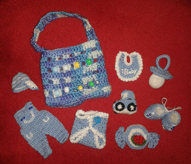 Handgehäkelte Babyartikel in Blau und Weiß auf rotem Hintergrund: Tasche mit Perlen, Strampler, Windelüberzug, Mütze, Lätzchen, Schnuller, Auto, Schühchen und Bonbon-Spielzeug.