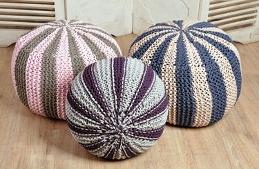 Drei gestrickte, gestreifte Poufs in rosa, grau, navyblau, beige und lila auf Holzboden.