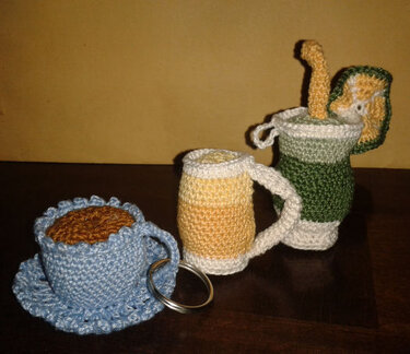 Drei gehäkelte Miniaturgefäße: blaue Tasse mit Untertasse, gelber Bierkrug und grüner Bierseidel mit Deckel auf dunkler Oberfläche; Metallring sichtbar.