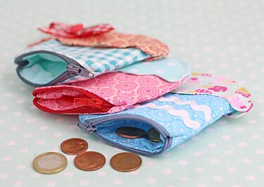 Draufsicht auf drei kleine, bunte Stoffgeldbörsen mit Reißverschlüssen auf hellgrünem, gepunktetem Hintergrund; eine enthält Münzen.