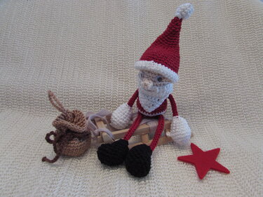 Gehäkelter Weihnachtsmann in Rot und Weiß auf kleinem Holzschlitten, daneben ein brauner Sack und ein roter Stern.