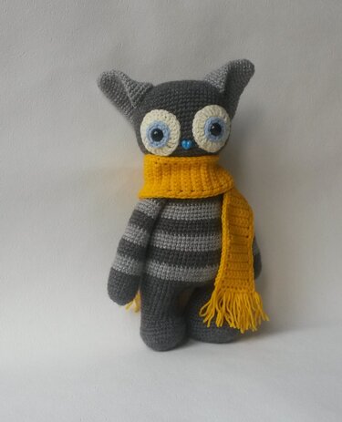 Gehäkeltes gestreiftes Amigurumi mit großen Augen und gelbem Schal vor hellem Hintergrund.