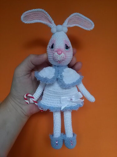 Gehäkeltes weiß-rosa Amigurumi-Häschen mit Kleid und Zuckerstange, gehalten von einer Hand vor orangefarbenem Hintergrund