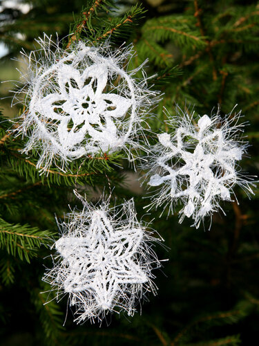 Drei weiße, flauschige gehäkelte Schneeflocken und Sterne an grünen Tannenzweigen