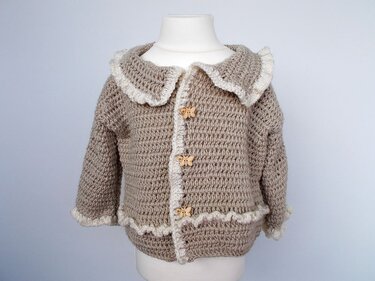 Beige gehäkelte Babyjacke mit weißer Rüsche und Schmetterlingsknöpfen auf Schaufensterpuppe