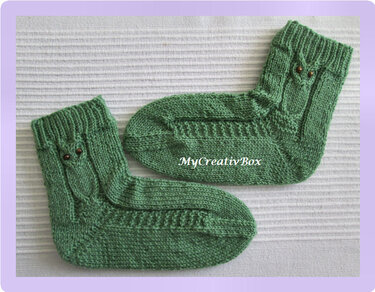 Paar grüne Babysocken mit Eulen-Zopfmuster und Knopfaugen auf hellem Hintergrund