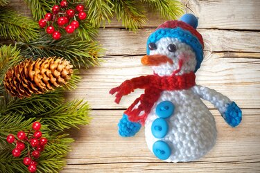 Gehäkelter Schneemann mit Mütze und Schal neben Tannengrün und Zapfen auf Holz