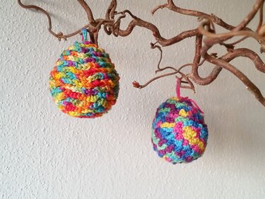 Zwei bunte, gehäkelte Ostereier an geschwungenen Zweigen vor heller Wand.