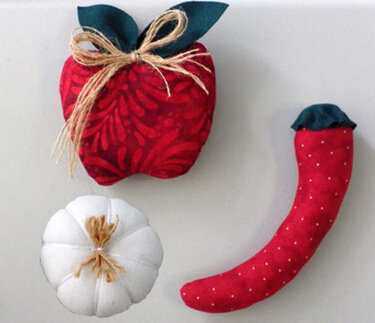 Drei genähte Stoffobjekte: roter Apfel mit Blättern und Bastband, weißer Kürbis und rote Chili mit Punkten.