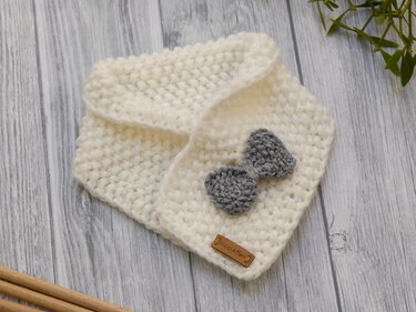 Weißes gestricktes Baby-Halstuch im Perlmuster mit grauer Schleife auf hellem Holzuntergrund; Stricknadeln und Zweig am Rand.