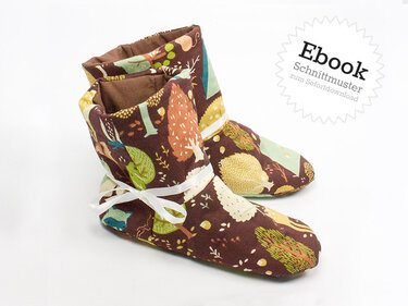 Braune Kinderhausschuhe mit buntem Wald- und Baummuster, weißes Band, vor hellem Hintergrund.