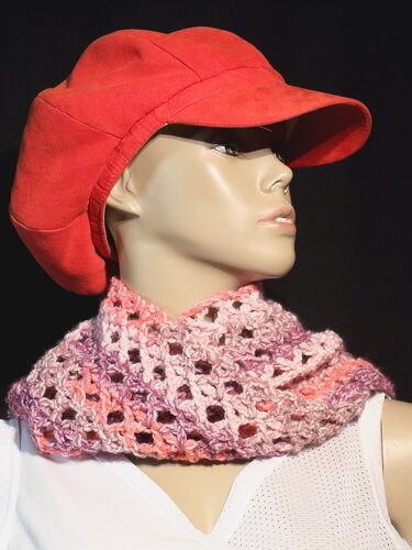 Schaufensterpuppe trägt gehäkelten Loop-Schal in Rosa, Lila und Orange sowie eine rote Kappe vor dunklem Hintergrund.