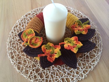 Weiße Stumpenkerze, umgeben von einem gehäkelten Kranz in gestreiften Herbsttönen, verziert mit dunkelbraunen Blättern und bunten Blüten auf einem geflochtenen Untersetzer.