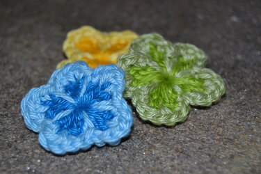 Drei gehäkelte Blumen‑Applikationen in Blau, Grün und Gelb auf dunklem Hintergrund.