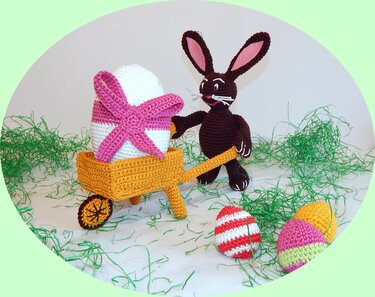 Stehender gehäkelter Hase mit rosa Innenohren neben gelber Schubkarre, großem Osterei und mehreren kleinen gehäkelten Eiern