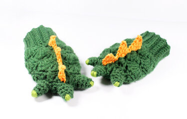 Grüne gehäkelte Drachenhandschuhe mit orangen Zacken und hellgrünen Krallenspitzen auf hellem Hintergrund.