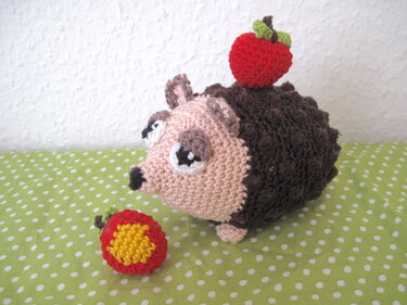 Gehäkelter Igel-Amigurumi in Braun und Rosa mit zwei gehäkelten Äpfeln auf grünem, gepunktetem Stoff