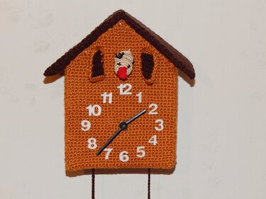 Handgehäkelte Kuckucksuhr in Hausform mit braunem Dach, orangefarbenem Körper, gehäkeltem Vogelkopf und weißen Ziffern