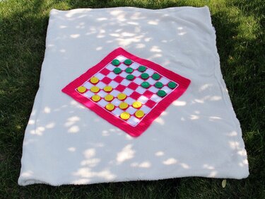 Buntes gehäkeltes Damebrett mit gelben und grünen Spielfiguren auf heller Decke im Gras