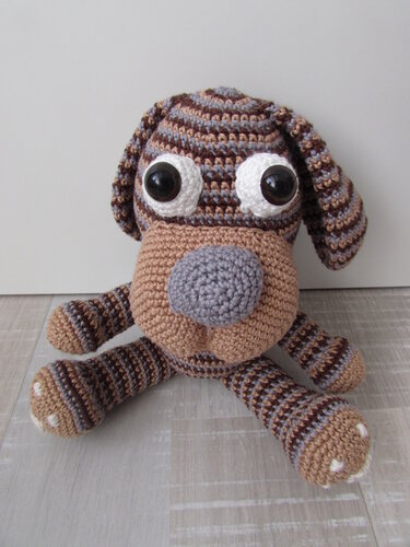 Gestreifter Amigurumi‑Hund in Braun-, Beige- und Grautönen, sitzend mit großen Augen und Schlappohren.