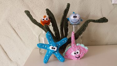 Mehrere gehäkelte Meerestiere (Fische, Seestern, Rochen) mit gehäkelten Wasserpflanzen in Glasflaschen.