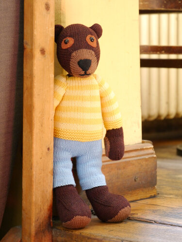 Hand-knitted brown teddy bear wearing a yellow-and-white striped sweater and light blue trousers, leaning against a wooden post.