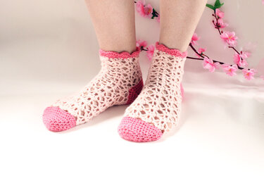 Kurze, luftige gehäkelte Socken in hellrosa mit pinker Ferse und Zehe sowie gezacktem Bündchen, auf hellem Hintergrund mit rosa Blütenzweig
