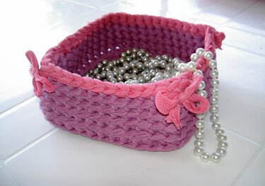 Gehäkelter Aufbewahrungskorb in Lila und Pink, gefüllt mit Perlen; Perlenkette hängt über den Rand.