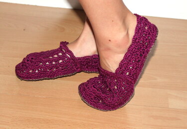 Füße mit gehäkelten auberginefarbenen Ballerina-Hausschuhen auf hellem Holzfußboden