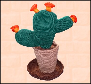 Gehäkelter Kaktus mit drei orangen Blüten in einem beigen Topf auf einer braunen Untertasse vor einer hellen Ziegelwand.