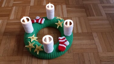Grüner gehäkelter Adventskranz mit vier weißen Kerzenüberzügen, gelben Sternen und rot-weiß gestreiften Mini-Strümpfen auf Parkettboden