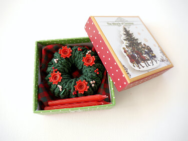 Gehäkelter dunkelgrüner Adventskranz mit roten Stern-Kerzenhaltern und Stabkerzen in offener Geschenkbox auf kariertem Stoff.