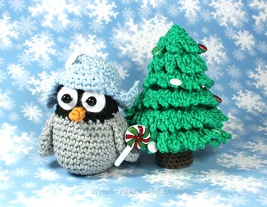 Gehäkelter Pinguin mit blauer Mütze neben kleinem gehäkeltem Weihnachtsbaum und Lolli vor Schneeflockenhintergrund