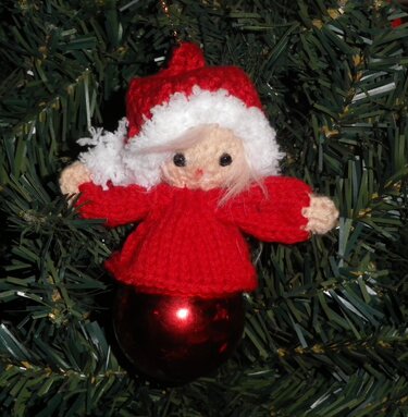 Gestrickter roter Amigurumi-Engel mit weißem Mützensaum auf roter Kugel vor Tannengrün