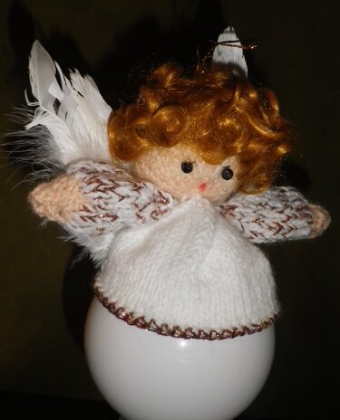 Gestrickter Engel mit rötlich-braunem Lockenhaar, schwarzen Augen, weißem Strickkleid mit goldenen Glitzerdetails und flauschigen weißen Federflügeln.