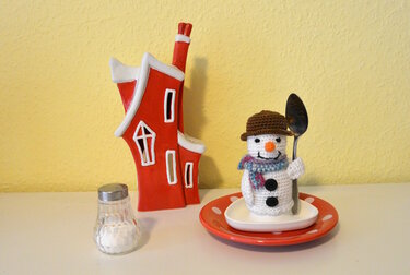 Gehäkelter Schneemann mit braunem Hut und buntem Schal auf Teller, neben rotem Dekohäuschen und Salzstreuer, hält einen Löffel.