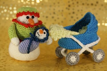 Amigurumi-Schneemann hält ein Baby, daneben ein blauer gehäkelter Kinderwagen vor goldenem Sternenhintergrund.