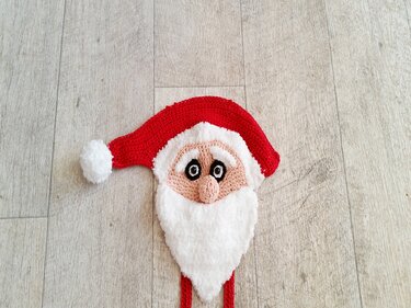 Gehäkelter Weihnachtsmannkopf mit roter Mütze und weißem Bart auf hellem Holzboden