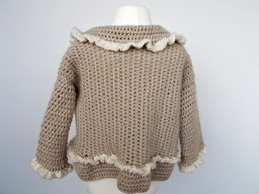 Rückansicht einer beigen gehäkelten Babyjacke mit cremefarbenen Rüschen an Kragen, Ärmeln und Saum auf einer Schaufensterpuppe.