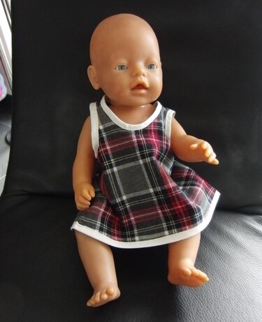 Baby doll wearing grey, red and white plaid sleeveless dress with white binding, seated on a dark surface.