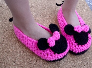 Child wearing bright pink crocheted slip-on slippers with black mouse-shaped appliqués and small pink bows