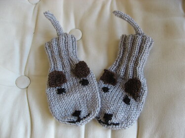 Zwei hellgraue, gestrickte Baby-Fäustlinge mit gestickten Hundegesichtern und braunen Ohren auf gepolsterter hellbeiger Oberfläche.