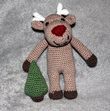 Handmade crocheted amigurumi reindeer with red nose holding a small green tree