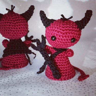 Zwei gehäkelte Amigurumi-Teufel, rot mit schwarzen Hörnern und Schwanz, einer hält einen Dreizack.
