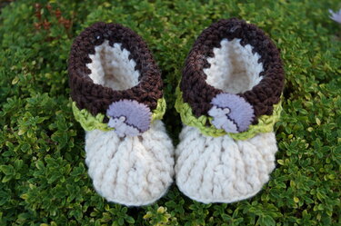 Paar handgehäkelter Babyschuhe in Creme mit braunem Bündchen und Igel-Knöpfen auf grünem Laub