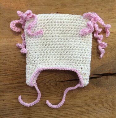 Gehäkelte cremefarbene Babymütze mit rosa Kante, Ohrenklappen, Bindebändern und spiralförmigen Dekorationen