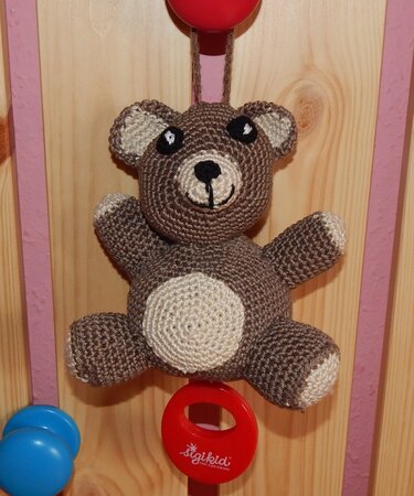 Gehäkelter brauner Teddybär mit beigem Bauch, aufgehängt an einem roten Ring vor Holzhintergrund.