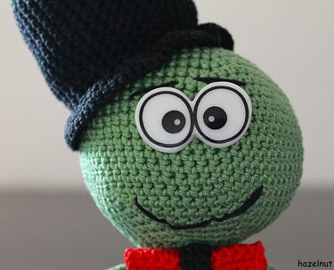 Gehäkelte grüne Amigurumi-Figur mit schwarzem Zylinder und roter Fliege, Nahaufnahme
