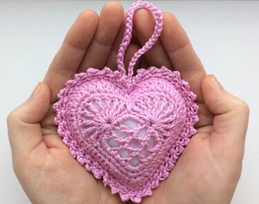 Hände, die ein rosafarbenes gehäkeltes Herz mit Aufhängeschlaufe halten