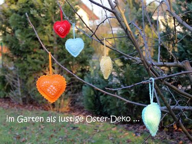 Mehrfarbige gehäkelte Herzen und blattförmige Anhänger hängen an Zweigen im Garten.